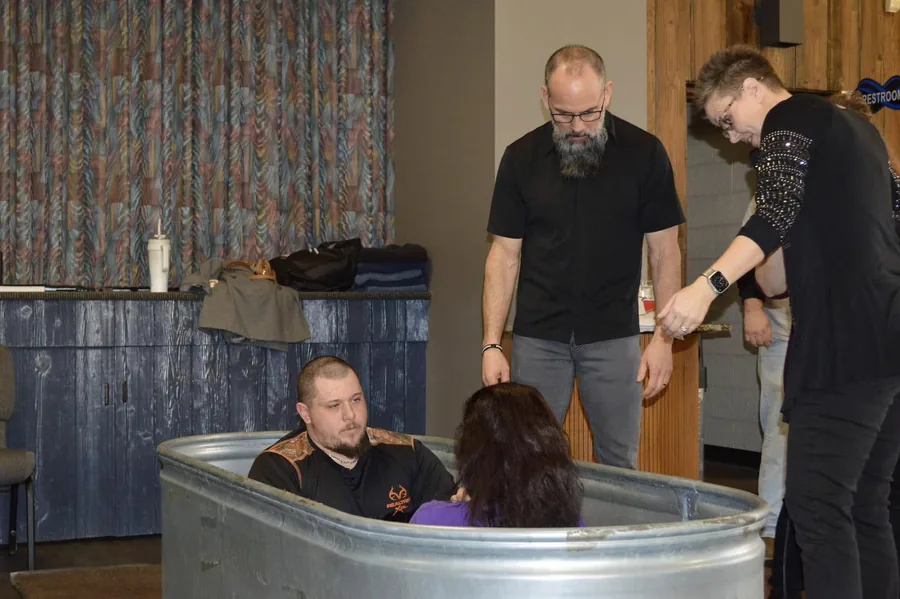 A man being baptized by immersion in a stock tank, supported by two Rush Church leaders