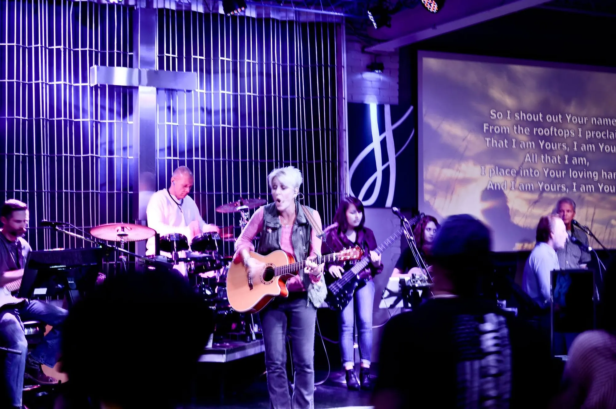 The Rush Church worship band leading a song on stage under purple and magenta lighting, with lyrics projected on the screen beside a large cross